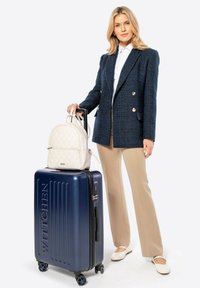 Marineblauer Hartschalenkoffer mit Rillen, schwarzem Reißverschluss und vier Rollen; cremefarbene gesteppte Backpack; Modell in strukturiertem blauen Blazer und beigen Hosen.