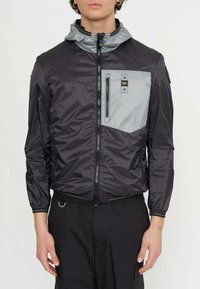 Giacca leggera nera e grigia con zip frontale, cappuccio e tasca sul petto. Presenta materiali a contrasto e polsini elasticizzati.