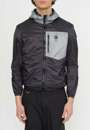Giacca leggera nera e grigia con zip frontale, cappuccio e tasca sul petto. Presenta materiali a contrasto e polsini elasticizzati.