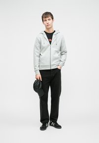 Hanorac gri deschis cu fermoar și glugă cu șnur, tricou grafic negru, blugi negri și adidași negri cu dungi albe. Ține în mână o șapcă neagră.