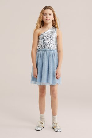 O fată tânără stă în picioare purtând un top argintiu cu paiete, cu un umăr gol, o fustă plisată bleu deschis și pantofi argintii cu șosete.