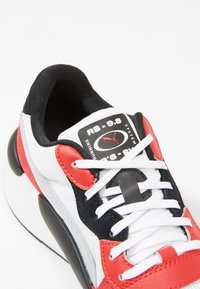 Red, white, and black sneaker with a fabric and leather upper, featuring a prominent logo, laces, and padded collar. Round sole.