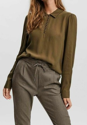Vrouw draagt een olijfgroene blouse met lange mouwen en stofbedekte knopen en een bruine geruite broek met hoge taille en een strik in de taille.