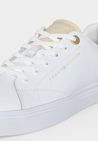 Λευκό αθλητικό παπούτσι Tommy Hilfiger με λευκά κορδόνια, χρυσά μεταλλικά κουμπώματα, μπεζ υφασμάτινη γλώσσα και χρυσό λογότυπο Tommy Hilfiger στο πλάι.