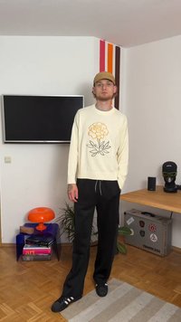 Cremfarbener Strickpullover mit orangefarbenem Blumenmuster, kombiniert mit schwarzen Hosen und schwarzen Sneakers. Lässiger, lockerer Schnitt und minimale Details.