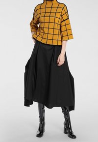 Żółta musztardowa bluzka w kratę z krótkim stojącym kołnierzykiem, zestawiona z czarną, sięgającą kolan, rozkloszowaną spódnicą. Całość dopełniają błyszczące czarne kozaki za kolano.