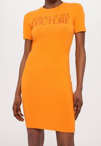 Orange t-shirt klänning med en fitted silhuett, korta ärmar och tryckt text "VERSACE JEANS COUTURE" i en kontrasterande färg. Slätt bomullstyg.