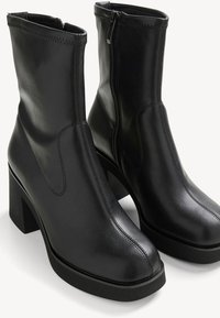 Paire de bottines en cuir noir avec talons blocs et fermetures éclair latérales sur fond blanc.