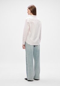 Svijetlo ružičasta košulja sa prugama i ovratnikom na dugme, uparena sa širokim svijetlo plavim trapericama koje imaju detalj dugmeta sa strane.