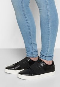 Černé kožené sneakersy s bílou podrážkou, s elastickými pásky přes přední část a hladkým povrchem.Částečně viditelné modré džíny.