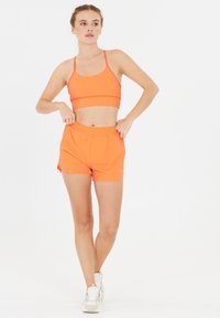 Ljusorange idrottstopp med tunna axelband och matchande shorts, med resår i midjan. Slät, lättviktsmaterial, lämplig för träning.
