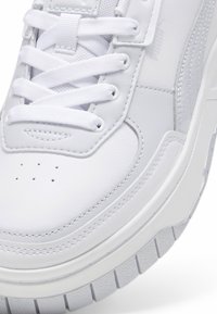 Tenis din piele albă cu accente textile, șireturi plate, cutie de vârf perforată și o talpă din cauciuc texturată. Prezintă detalii subtile de branding.