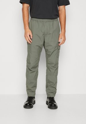 Mann trägt olivgrüne Cargohosen und schwarze Schnürstiefel, steht vor einem einfarbigen hellen Hintergrund.
