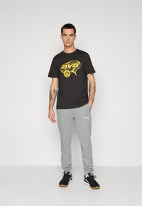 Mand iført sort BVB-logo t-shirt, grå Puma joggingbukser og sorte sneakers står mod en plain lys baggrund med afslappede hænder.