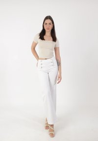 Mujer de pie con una mano en la cadera, vestida con una blusa beige ajustada, pantalones blancos de tiro alto con botones, sandalias beige y un tatuaje visible en el brazo.