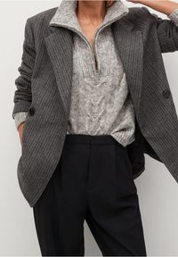 Šedé sako s proužky nošené přes světle šedý svetr s vysokým límcem a kabelovým vzorem. Kombinováno s tmavými, na míru šitými kalhotami.