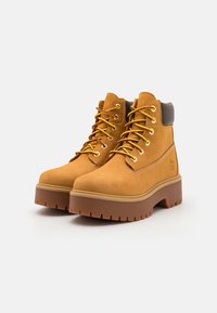 Botas de cano curto em couro castanho claro com atacadores amarelos, colarinho acolchoado e sola de borracha robusta com uma plataforma grossa e padrão de ranhura profundo.