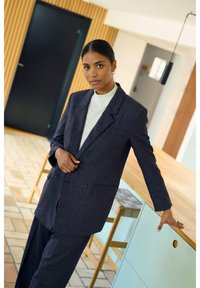Blazer sartorial blu scuro con rever a punta, chiusura a doppiopetto e tasche laterali, abbinato a pantaloni coordinati. Tessuto liscio.