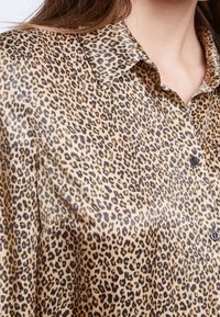 Luipaardprint overhemd in een lichte, zijden stof. Het shirt heeft een klassieke kraag, een knoopsluiting aan de voorkant en is voornamelijk in een tan kleur met zwarte vlekken.