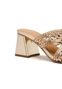 Sandale à talon dorée en tissu tressé avec un talon bloc, ornée d'un accent miroir. La partie supérieure présente un design texturé et tressé.
