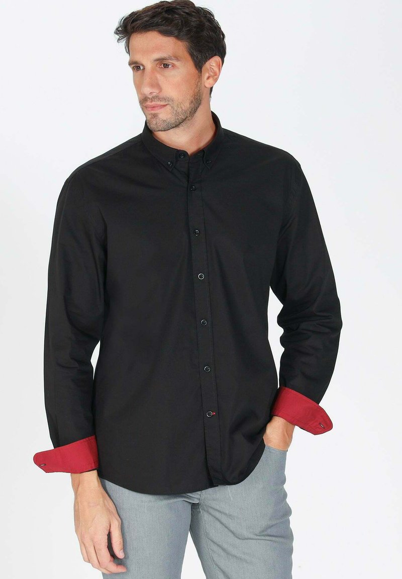 Chemise noire à manches longues avec un col boutonné, présentant des poignets contrastants rouges et des boutons noirs, accompagnée d'une coupe droite.