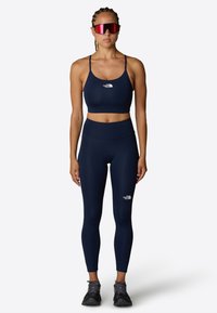 Laivaston urheilullinen crop top ja leggings, valmistettu joustavasta materiaalista, sisältävät valkoisia logo-yksityiskohtia. Mustat urheilujalkineet täydentävät asua.