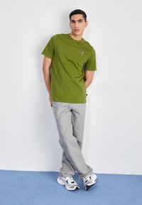 Grøn bomulds T-shirt med korte ærmer, prydet med et sort logo. Kombineret med lysegrå bukser og sort-hvide sneakers.