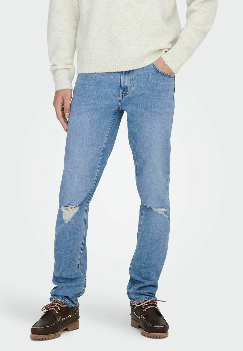 Jeans de mezclilla de un azul claro con corte ajustado, que presentan detalles desgastados en las rodillas y un diseño estándar de cinco bolsillos. Combinados con zapatos marrones.
