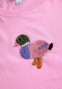 Roza bombažna majica s potiskom račke iz perlic v zelenih, vijoličnih, roza, črnih in oranžnih odtenkih. Teksturirano delo s perlami ustvarja globino.