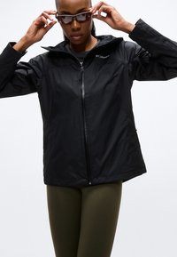 Γυναίκα που προσαρμόζει τα γυαλιά ηλίου, φοράει μαύρο τζάκετ με φερμουάρ Columbia και χακί κολάν, σε λευκό φόντο.