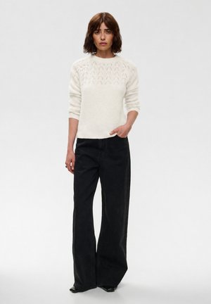 Donna in piedi che indossa un maglione bianco lavorato a maglia con una pezza a motivo e pantaloni neri a gamba larga, su uno sfondo chiaro uniforme.
