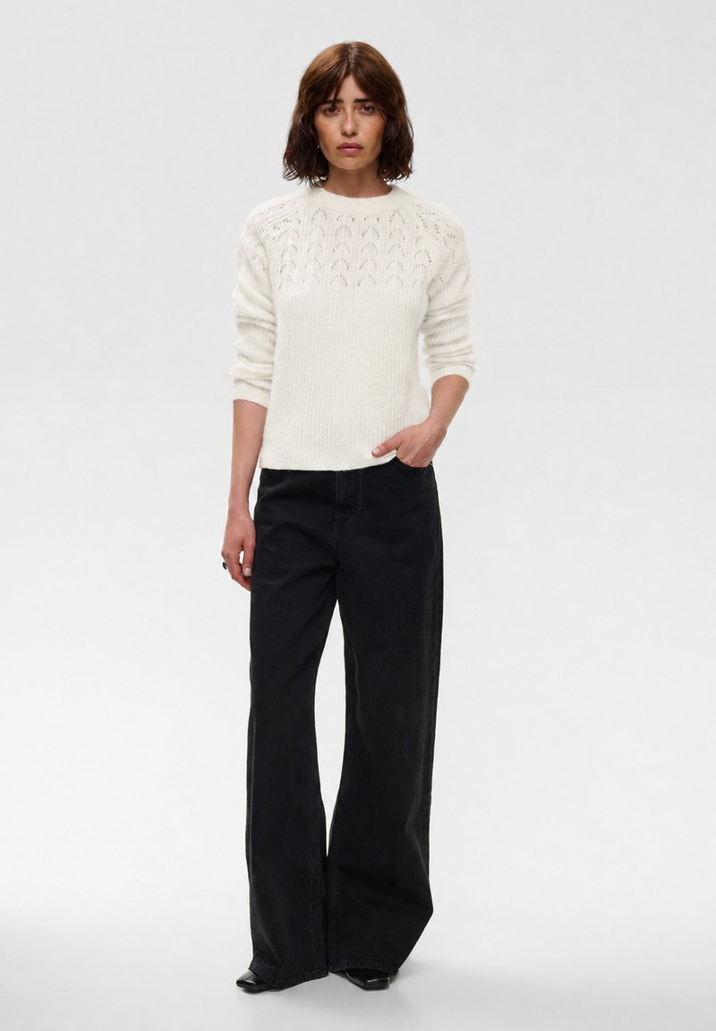 Femme debout portant un pull en maille blanc avec un empiècement à motifs et un pantalon noir à jambes larges, sur un fond clair uni.