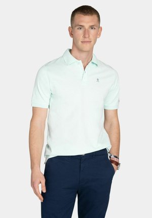 Młody mężczyzna w jasnym miętowym polo i ciemnoniebieskich spodniach, stojący z jedną ręką w kieszeni na jednolitym tle.