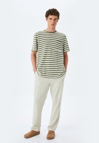 Randig t-shirt i olivgrön och vit med rund halsringning och ficka, kombinerad med ljusbeiga byxor och bruna slip-on skor.