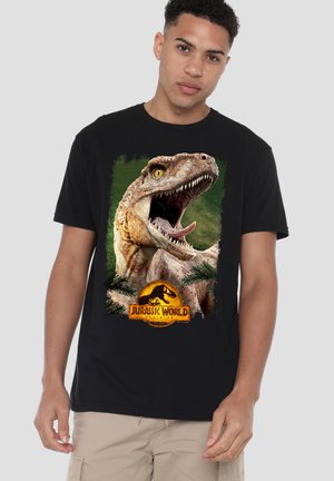 Junger Mann trägt schwarzes T-Shirt mit geöffnetem Dinosaurier-Mund und Jurassic World Dominion-Logo vorne vor einfarbigem Hintergrund.