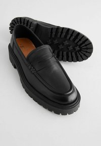 Black leather loafers with a thick, rugged rubber sole, featuring a rounded toe, subtle top stitching, and a classic penny strap detail.