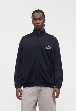 Giovane uomo con capelli intrecciati indossa una giacca nera Carhartt WIP con zip e pantaloni grigio chiaro, in piedi davanti a uno sfondo bianco uniforme.