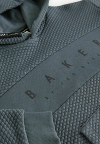 Felpa grigia scura con texture trapuntata sulle spalle e un pannello liscio con la scritta "BAKER by TED BAKER" in nero.