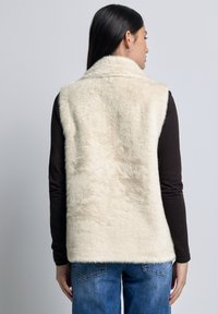 Gilet in finta pelliccia color crema con una consistenza morbida, caratterizzato da un colletto e una vestibilità comoda. Indossato sopra una maglietta a maniche lunghe marrone e jeans blu.