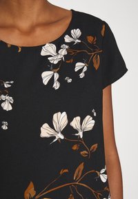 Haut noir à manches courtes avec des branches marron et un motif floral blanc, porté par une personne à la peau de ton moyen-foncé.