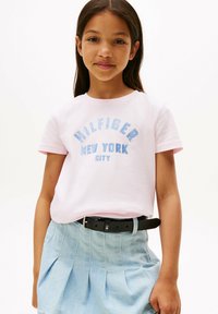 Camiseta rosa claro con el texto azul brillante "HILFIGER NEW YORK CITY", combinada con una falda de mezclilla clara y un cinturón negro. Mangas cortas, cuello redondo.