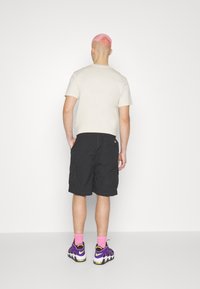 T-shirt beige clair pour homme à col rond, short cargo noir et chaussures de sport violettes avec chaussettes roses, vus de dos.