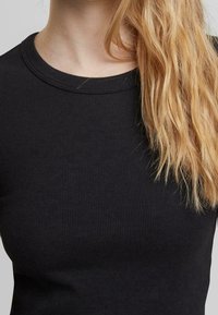 Cheveux blonds tombant sur l'épaule droite, portant un haut noir côtelé à col rond sur un fond neutre.