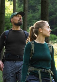 Kaksi vaeltajaa metsässä. Mies pukeutuu mustaan pitkähihainen paitaan ja harmaisiin kuvioituihin housuihin, kun taas naisella on yllään vihreä pitkähihainen paita ja housut.