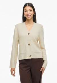 Žena se smije, odjevena u bež kardigan s gumbima i prednjim džepovima, u tamnosmešnim hlačama, stoji pred bijelom pozadinom.