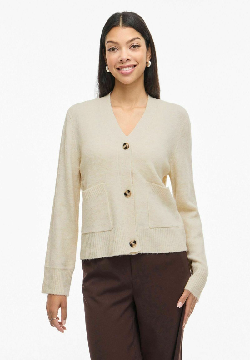 Žena se smije, odjevena u bež kardigan s gumbima i prednjim džepovima, u tamnosmešnim hlačama, stoji pred bijelom pozadinom.