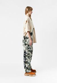 Mujer de pie de lado, vistiendo una blusa beige oversized y pantalones grises-negros con un gráfico de etiqueta de equipaje negra, con zapatos chunky marrones sobre un fondo blanco.