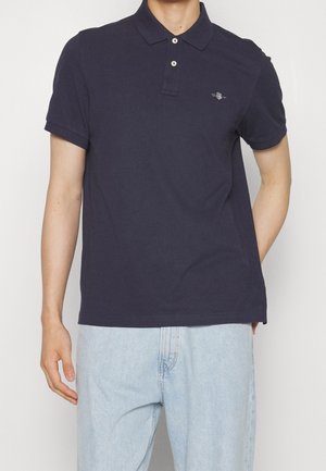 Homme portant un polo bleu marine à manches courtes avec deux boutons et un jean bleu clair, debout devant un fond uni.