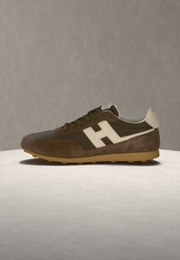 Baskets marron et beige avec une pièce blanche au talon, une semelle beige et un logo blanc en forme de lettre H sur le côté, placées sur une surface claire sur fond gris.