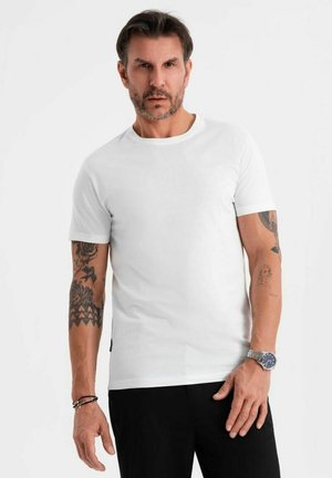 Camiseta blanca de manga corta con escote redondo, hecha de una tela suave, combinada con pantalones oscuros. Presenta tatuajes visibles en los brazos.